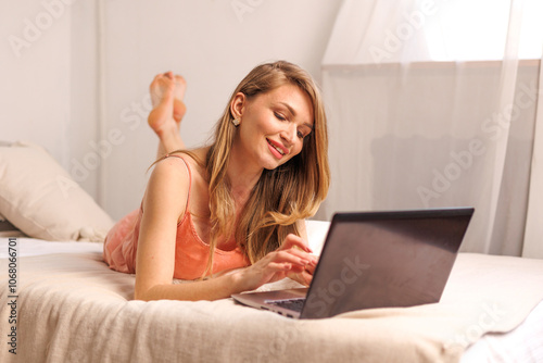 Young woman relaxing at home in the afternoon, using a laptop while holding a cup, enjoying her time in a cozy bedroom setting