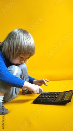 A small child counts on a calculator on a yellow background. Kid fingers press buttons and calculate studying math at home. Child makes arithmetic calculations with a calculator
