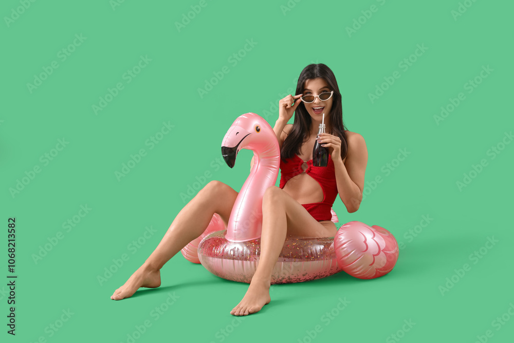 Young pretty woman in sunglasses and swimsuit sitting on flamingo inflatable ring with cocktail on green background Young pretty woman in sunglasses and swimsuit sitting on flamingo inflatable ring with cocktail on green background
