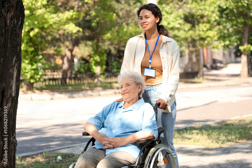 Young African-American female medical worker walking with elderly woman on wheelchair outdoors Young African-American female medical worker walking with elderly woman on wheelchair outdoors