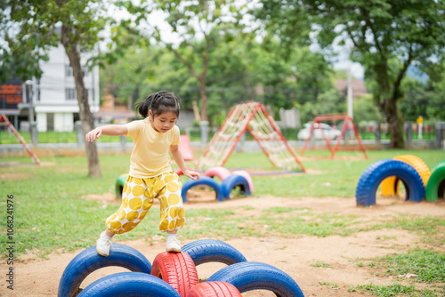 Asian young girl is playing in a park with a tire obstacle course. The park is filled with various play equipment, including swings and a slide. The atmosphere is lively and fun photo