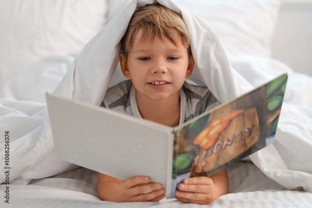 Naklejka premium Cute little happy boy in pajamas reading book on bed at home