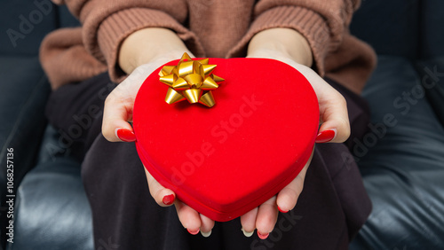 Asian woman wearing a brown shirt sit on black sofa and holding a red heart shaped gift box with a gold ribbon bow in hands. Red gift box. Presents and gift boxes for festivals and celebrations.