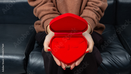 Asian woman sitting on black sofa and holding red heart shaped gift box with gold bow in hand. Gift box open lid, present and gift boxes for festival and celebration.