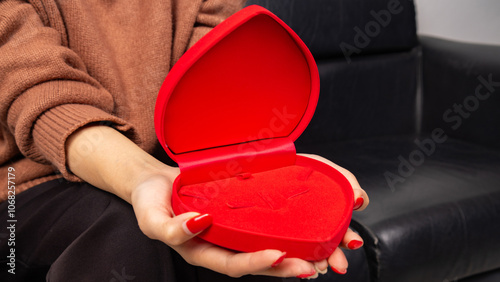 Asian woman sitting on black sofa and holding red heart shaped gift box with gold bow in hand. Gift box open lid, present and gift boxes for festival and celebration.