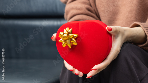 Asian woman wearing a brown shirt sit on black sofa and holding a red heart shaped gift box with a gold ribbon bow in hands. Red gift box. Presents and gift boxes for festivals and celebrations.