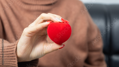 Asian woman wearing a brown shirt sit on black sofa and holding a little red heart shaped gift box in hands. Red gift box. Presents and gift boxes for festivals and celebrations.