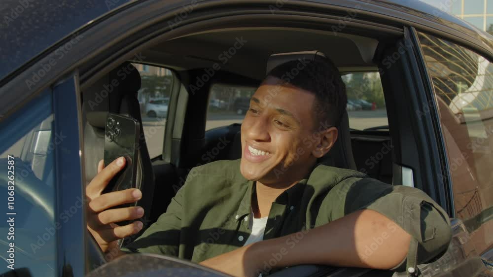 Portrait of attractive black male driver driving car in city street, video conferencing online with friend using cellphone , expressing positivity and friendliness during road trip.