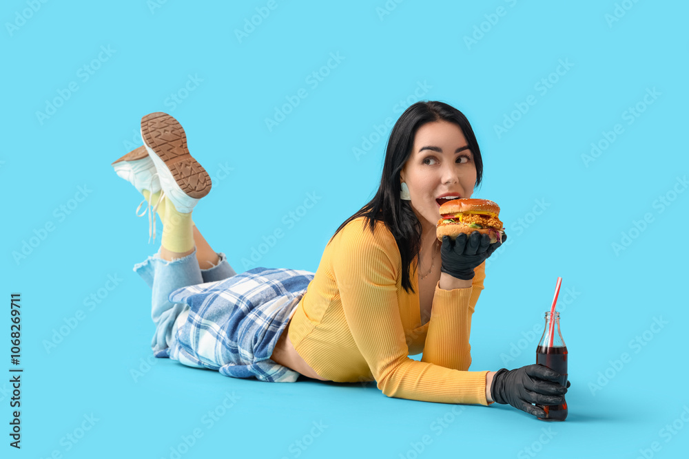 Beautiful young woman with tasty burger and bottle of cola lying on blue background Beautiful young woman with tasty burger and bottle of cola lying on blue background