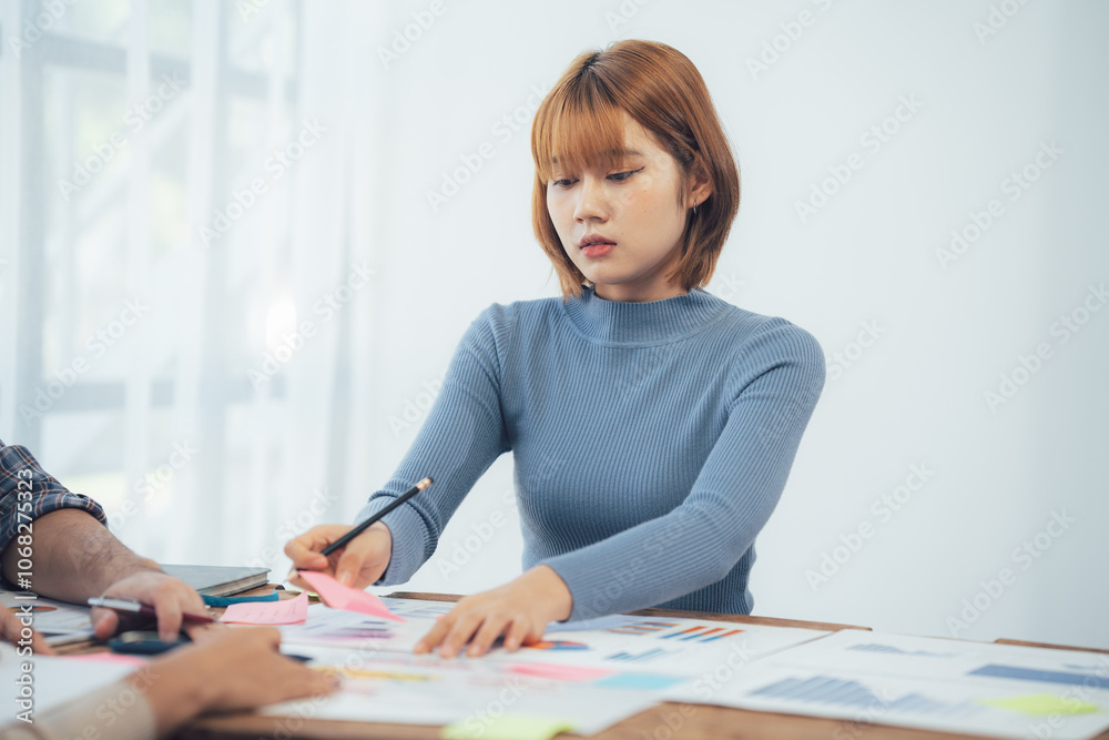 Focused Collaboration: A young Asian businesswoman intently reviews project plans during a collaborative meeting, showcasing dedication and strategic thinking. Focused Collaboration: A young Asian businesswoman intently reviews project plans during a collaborative meeting, showcasing dedication and strategic thinking.