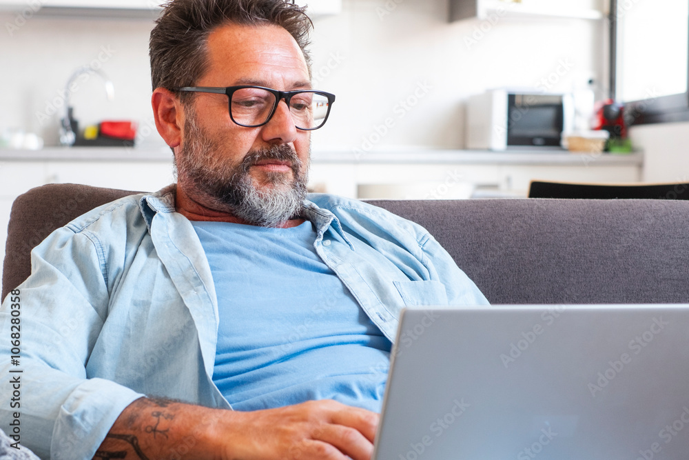 Middle-aged man focused on laptop screen while seated in his living room, glasses on, immersed in online work, digital projects, or exploring new career possibilities from home Middle-aged man focused on laptop screen while seated in his living room, glasses on, immersed in online work, digital projects, or exploring new career possibilities from home