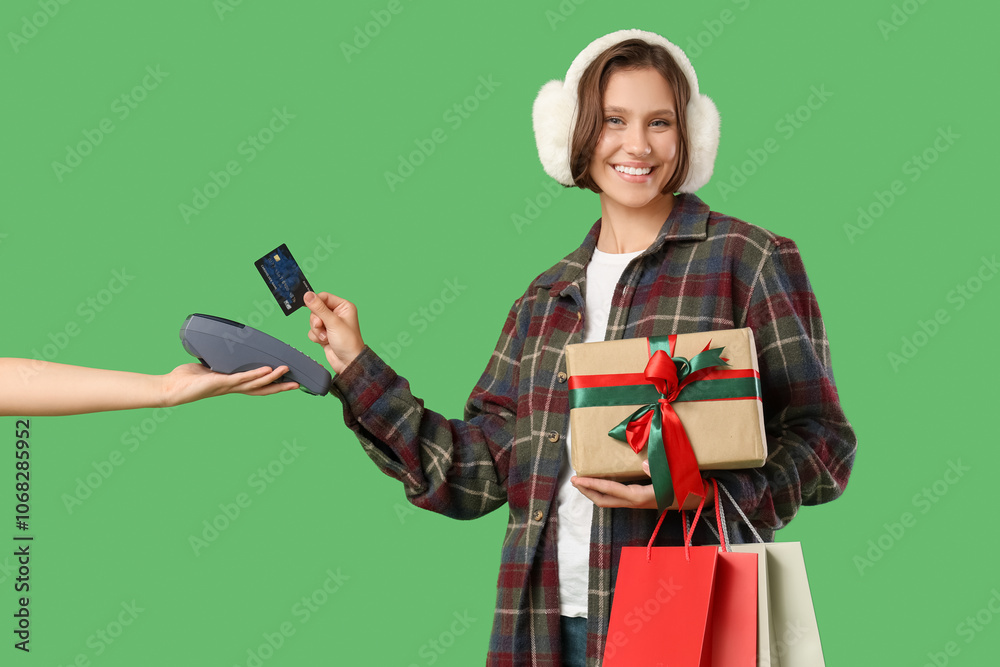 Beautiful young happy woman with shopping bags, gift box and credit card paying via payment terminal on green background. Christmas celebration Beautiful young happy woman with shopping bags, gift box and credit card paying via payment terminal on green background. Christmas celebration