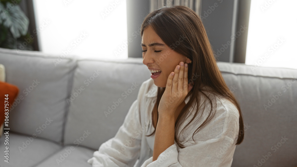 Young woman suffering indoors holding her cheek with a painful expression while sitting on a couch in her living room Young woman suffering indoors holding her cheek with a painful expression while sitting on a couch in her living room