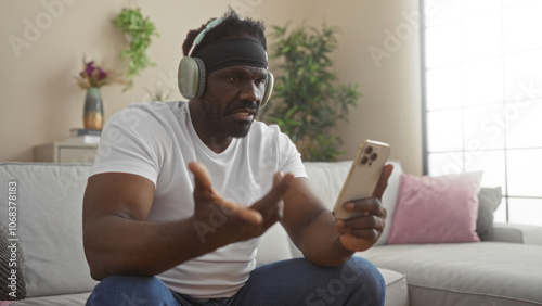 A handsome young african american man with a beard, wearing headphones, sits in a living room, holding a smartphone, appearing frustrated or confused during a video call in his apartment.