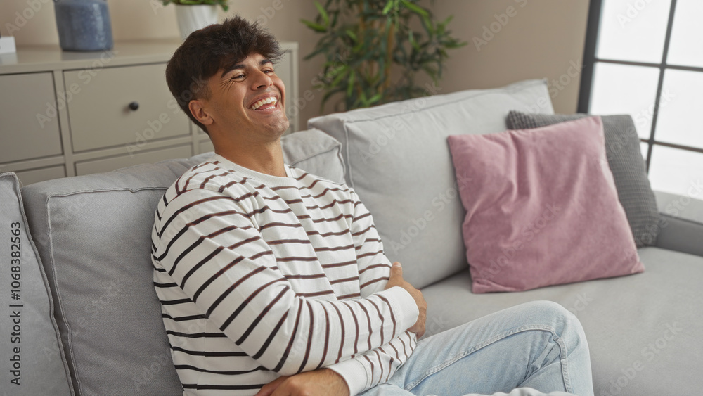 A handsome young hispanic man is laughing joyfully while sitting on a comfortable couch in a modern living room interior of an apartment. A handsome young hispanic man is laughing joyfully while sitting on a comfortable couch in a modern living room interior of an apartment.