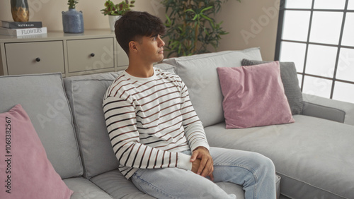 Young hispanic man sitting in a modern living room, reflecting on something, with a stylish interior featuring plants and cushions.