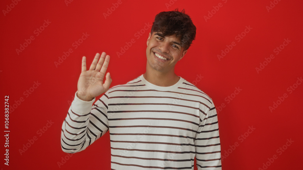 Naklejka premium Handsome young hispanic man smiling and waving in a striped shirt against an isolated red background.