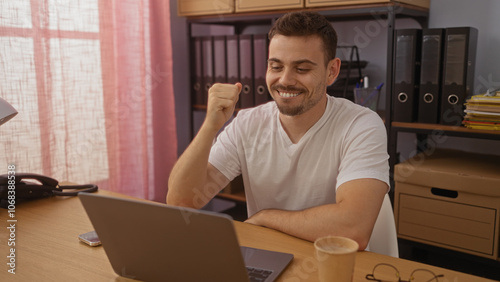 Smiling hispanic man with a beard in a white shirt working on a laptop in an office setting, celebrating a success with a fist pump, surrounded by office supplies and binders.