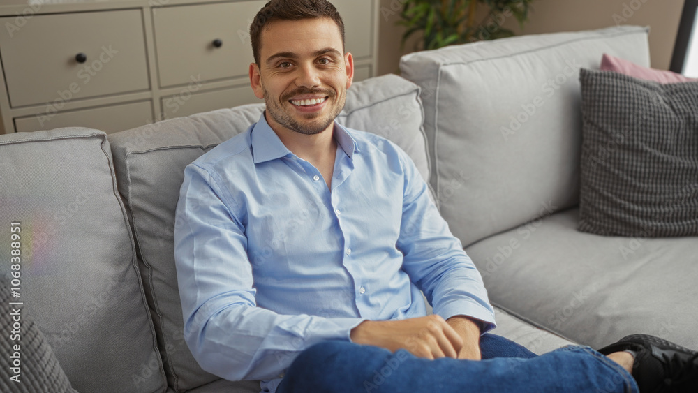 Handsome hispanic man with a beard smiling while sitting comfortably on a sofa in a modern living room at home. Handsome hispanic man with a beard smiling while sitting comfortably on a sofa in a modern living room at home.