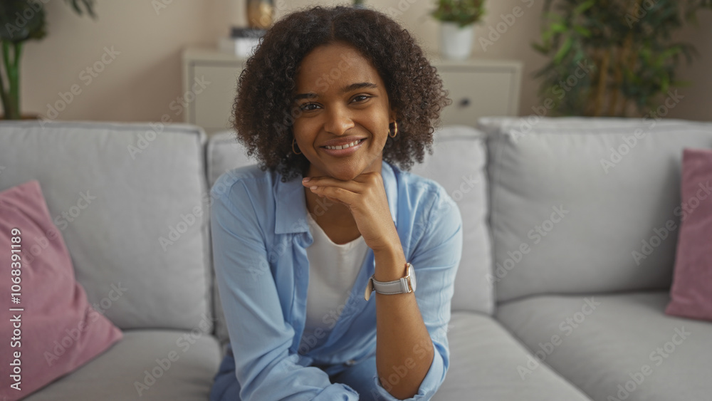 A beautiful young african american woman with curly hair smiles in a cozy living room, sitting comfortably on a couch in her apartment. A beautiful young african american woman with curly hair smiles in a cozy living room, sitting comfortably on a couch in her apartment.