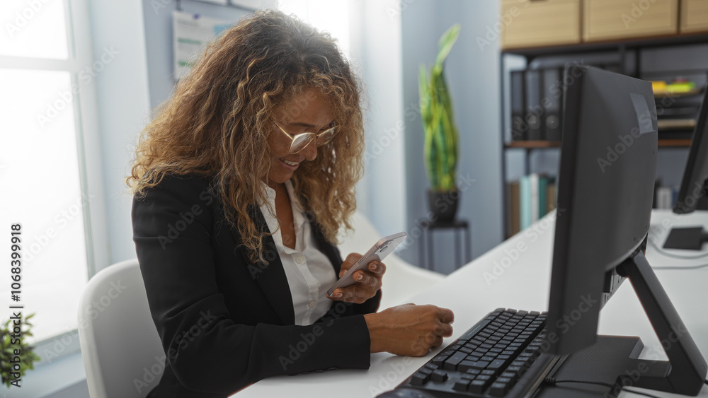 Obraz premium Woman working in an office, wearing glasses and using a smartphone at her desk, featuring a computer monitor, indoor plants, and shelves in the background.