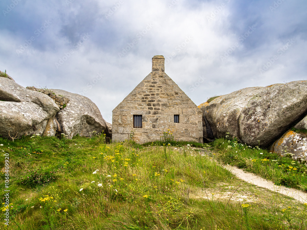 A small stone house nestled between two large boulders in Kerlouan, Brittany, in a grassy landscape under a cloudy sky. A small stone house nestled between two large boulders in Kerlouan, Brittany, in a grassy landscape under a cloudy sky.