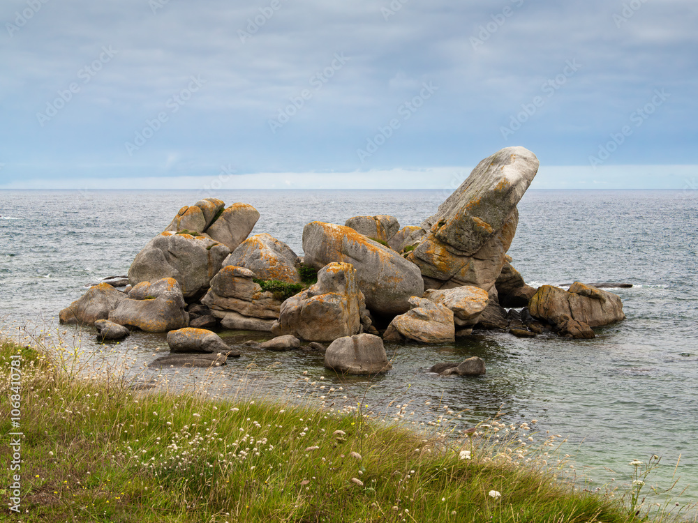 A rocky shoreline in Brittany with large boulders partially submerged in calm waters. The scene features a grassy area in the foreground, creating a serene coastal atmosphere. A rocky shoreline in Brittany with large boulders partially submerged in calm waters. The scene features a grassy area in the foreground, creating a serene coastal atmosphere.