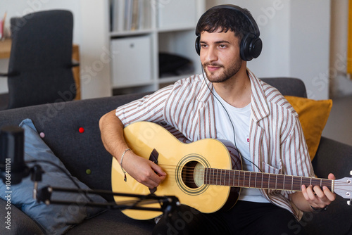 Arab young man in a home setting plays his acoustic guitar in front of a laptop, practicing his music with headphones on, illustrating a modern blend of digital tools and acoustic practice.
