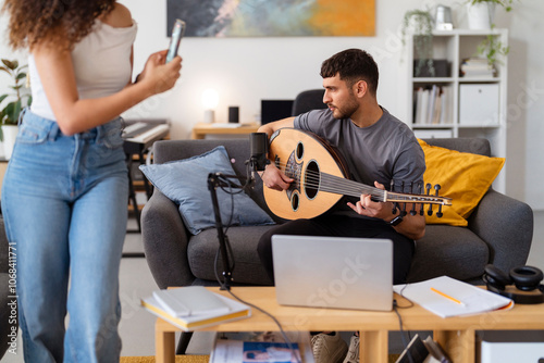 Arab woman records a musician playing the oud in a cozy living room, highlighting a creative and relaxed indoor session with a mix of traditional music and modern technology in a casual setting. photo