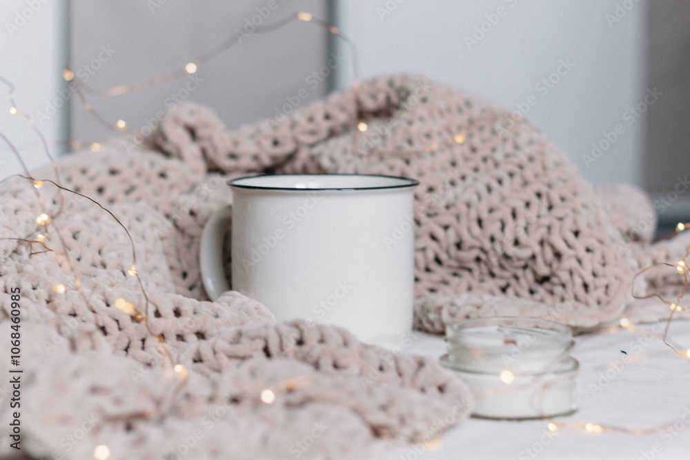 A white ceramic mug and a candle in a glass jar with a wooden wick stand among a knitted plush soft warm plaid. Warm comfort