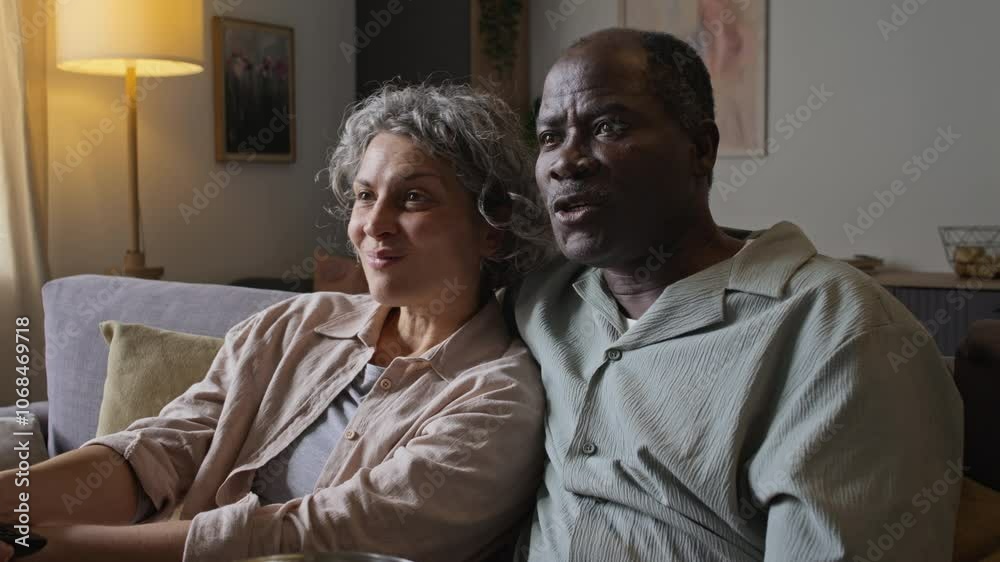 Tilt shot of happy diverse middle-aged married couple enjoying watching TV together at home, husband hugging wife from behind, spending leisure time together Tilt shot of happy diverse middle-aged married couple enjoying watching TV together at home, husband hugging wife from behind, spending leisure time together