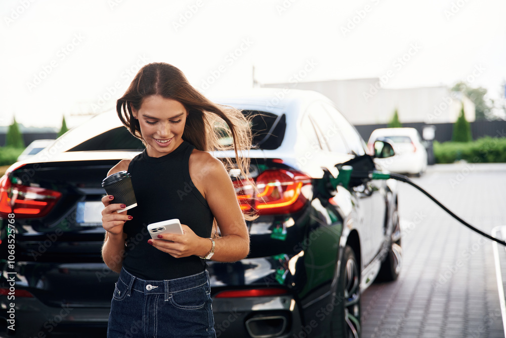 Waiting, holding smartphone. A young woman at a gas station with her car Waiting, holding smartphone. A young woman at a gas station with her car