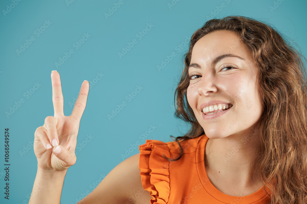 Portrait of a young woman on a blue background. She poses waving with her hand, showing a v with her fingers. Empowered and independent woman. Portrait of a young woman on a blue background. She poses waving with her hand, showing a v with her fingers. Empowered and independent woman.