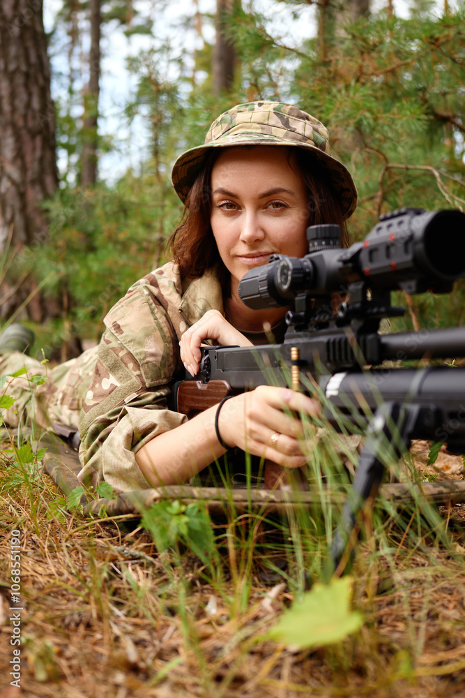 Fototapeta premium A focused female soldier lying in the forest, aiming with a sniper rifle during a training exercise in a natural setting