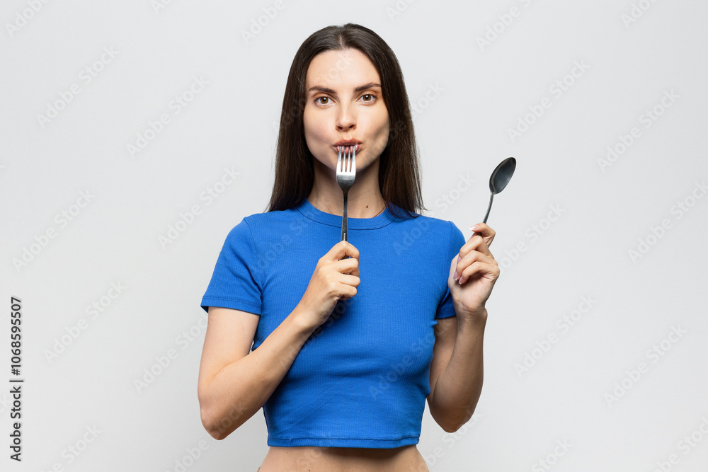 Cheerful young woman with blue top holding spoon and fork, starving and funny. On white background Cheerful young woman with blue top holding spoon and fork, starving and funny. On white background