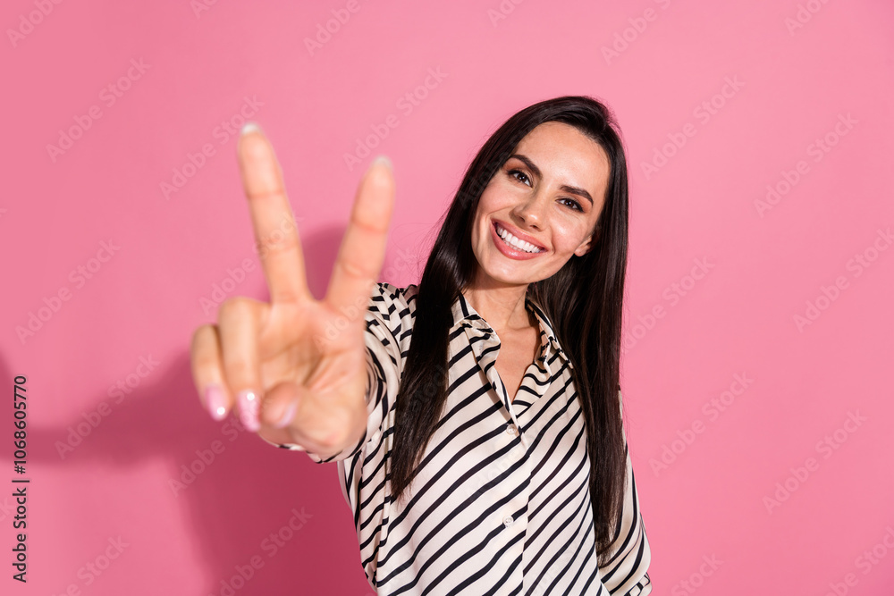 Young woman in striped shirt displays peace sign against vibrant pink background Young woman in striped shirt displays peace sign against vibrant pink background