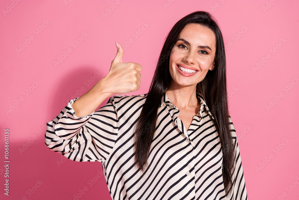 Confident young woman with brunette hair in striped shirt giving thumbs up against pink background
