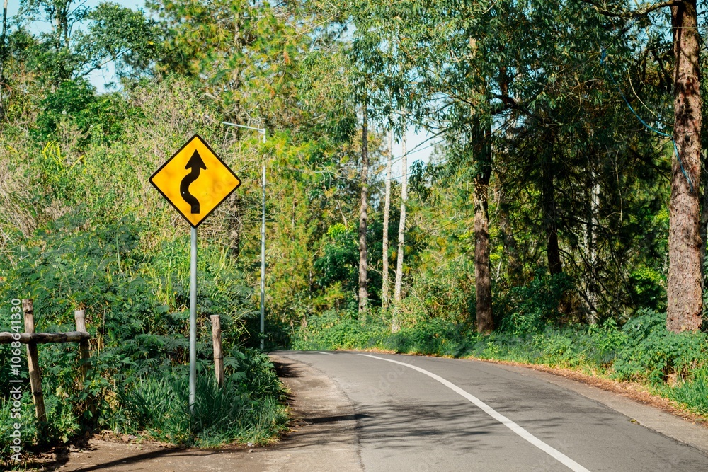 Winding road with yellow sign for a sharp curve through a forest. Traffic sign on mountain route. Winding road with yellow sign for a sharp curve through a forest. Traffic sign on mountain route.