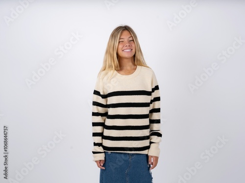 Coquettish caucasian teen girl wearing striped knitted sweater over white smiling happily, blinking at camera in a playful manner, flirting with you. photo