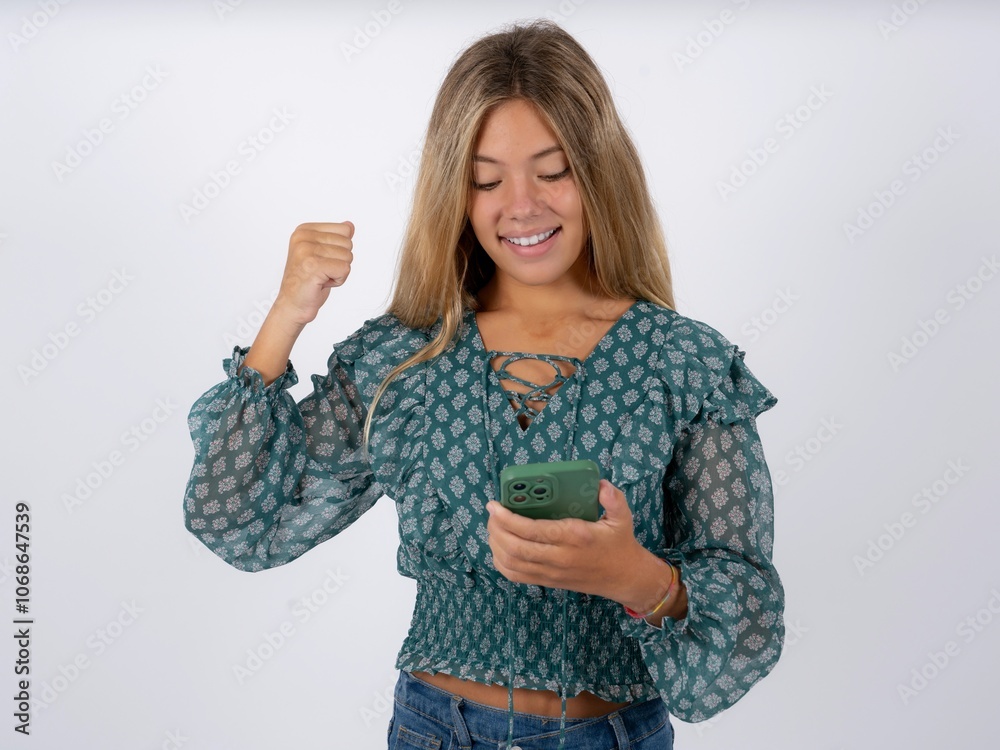 caucasian teen girl wearing green dress over white holding in hands cell and rising his fist up being excited after reading good news. caucasian teen girl wearing green dress over white holding in hands cell and rising his fist up being excited after reading good news.
