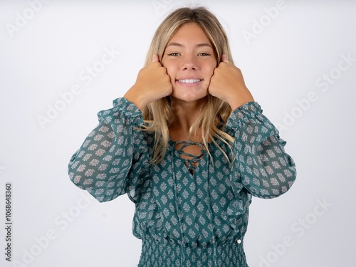 Happy caucasian teen girl wearing green dress over white keeps fists on cheeks smiles broadly and has positive expression being in good mood