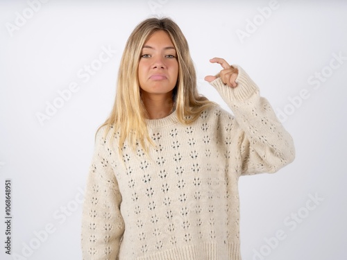 caucasian teen girl wearing knitted sweater over white purses lip and gestures with hand, shows something very little. photo