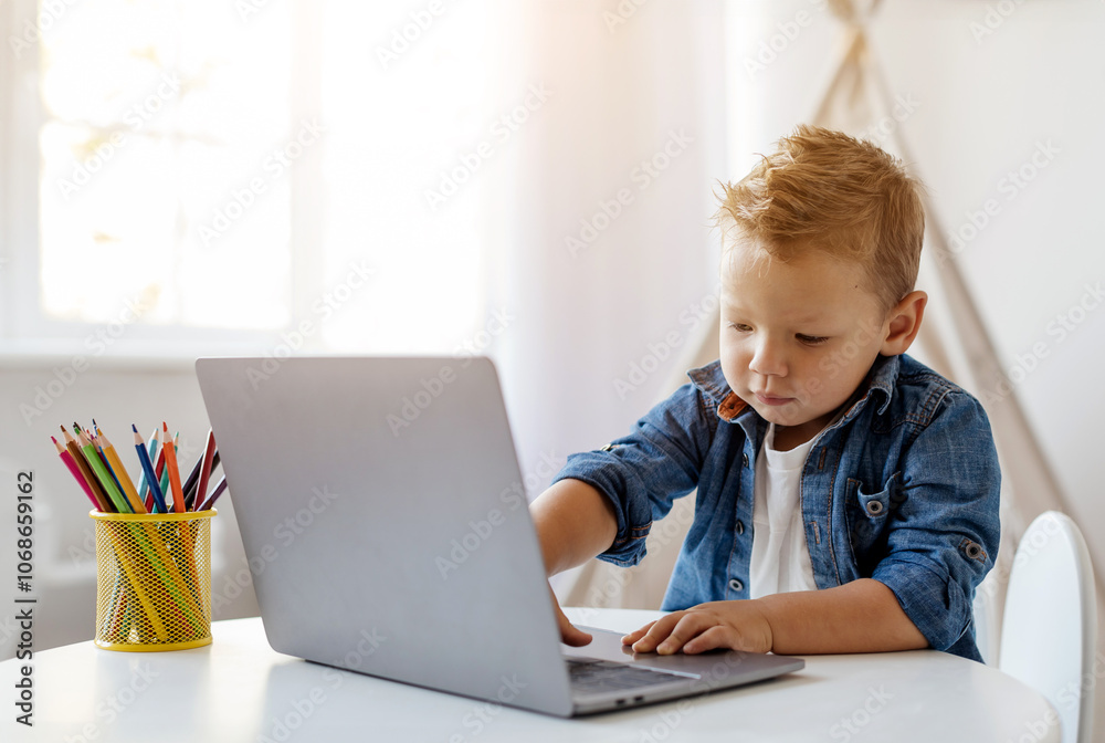 Portrait of little young boy focuses on his laptop at a table filled with colorful pencils in a well-lit room. The scene captures a moment of curiosity and learning. Portrait of little young boy focuses on his laptop at a table filled with colorful pencils in a well-lit room. The scene captures a moment of curiosity and learning.