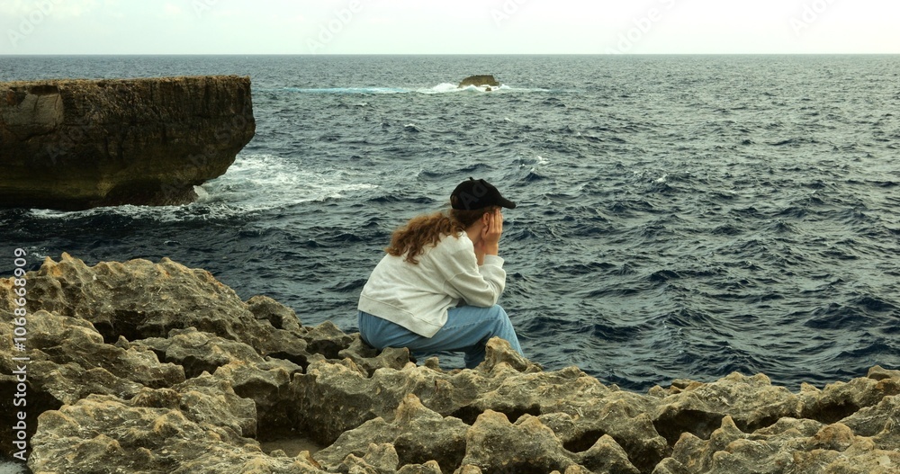Teenage girl contemplates rolling waves sitting alone on rock at sea resort. Upset teenager child rests on cliffy ocean beach during summer vacation Teenage girl contemplates rolling waves sitting alone on rock at sea resort. Upset teenager child rests on cliffy ocean beach during summer vacation