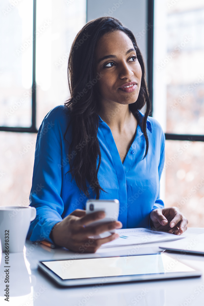 Thoughtful ethnic businesswoman using smartphone while sitting in office with devices Thoughtful ethnic businesswoman using smartphone while sitting in office with devices