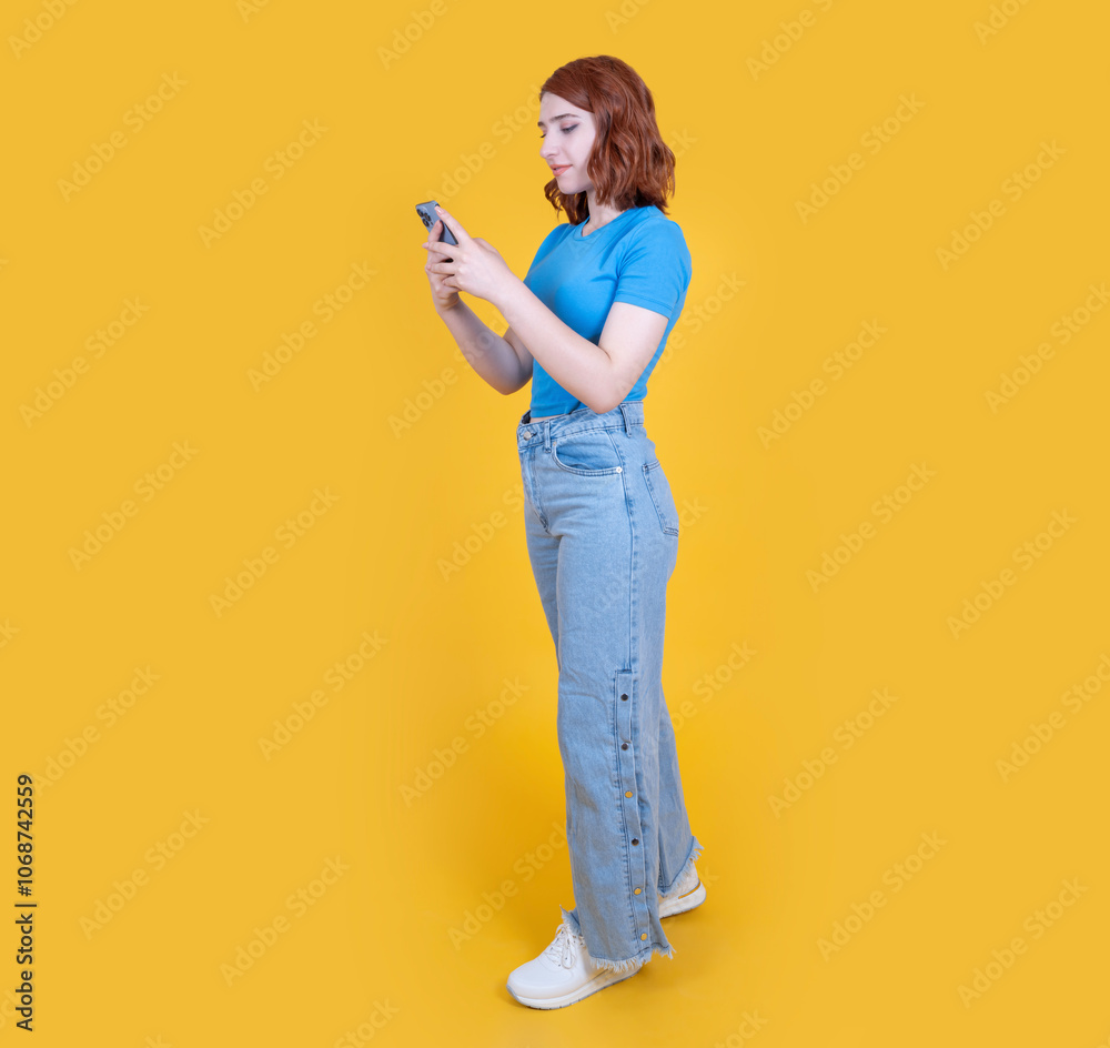Using smartphone, full body side view 20s young caucasian red bob hair woman using smartphone. Looking screen, holding typing message, shopping online, using trendy app over yellow background. Using smartphone, full body side view 20s young caucasian red bob hair woman using smartphone. Looking screen, holding typing message, shopping online, using trendy app over yellow background.