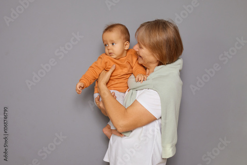 Mature woman holding her infant grandson with care smiling at grandchild feeling delighted and satisfied while playing indoors with newborn baby isolated over gray background