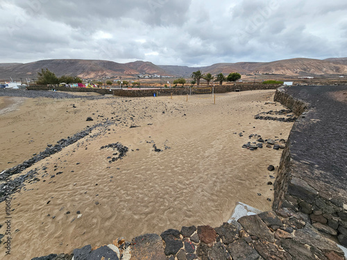 Plage de La Garita à Arrieta entourée de murs de pierre volcanique, avec des traces de roches dispersées et des montagnes arides en arrière-plan sous un ciel nuageux. photo