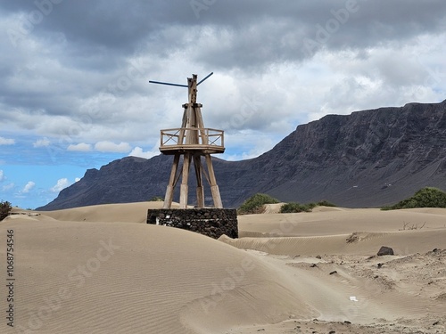 Une structure en bois ressemblant à un moulin, érigée sur un socle en pierre au milieu des dunes de sable, de Caleta de Famara, avec des montagnes en arrière-plan sous un ciel nuageux.