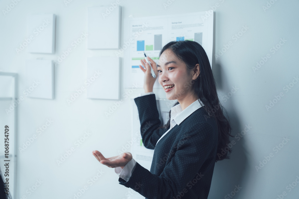 Confident Businesswoman Presenting Data: A young, smiling Asian businesswoman confidently presents data on a chart during a meeting.